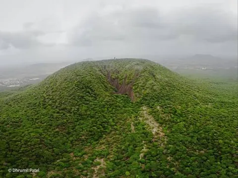 Aerial view of a rounded hill covered in green scrub after the monsoon, with an eroded gully on one side
