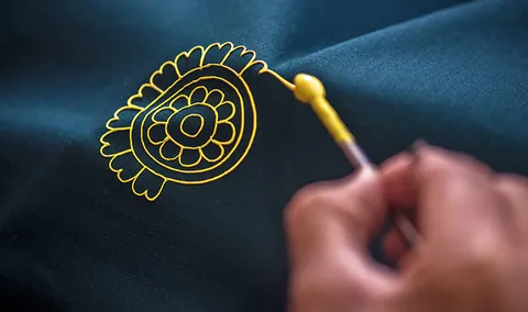 Close-up of a stylus drawing a yellow Rogan motif on dark cloth